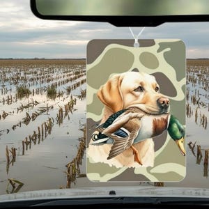 May include: Rectangular air freshener featuring a yellow Labrador Retriever holding a duck in its mouth. The background is a camouflage pattern. The air freshener hangs from a white string, with a field and sky visible in the background.