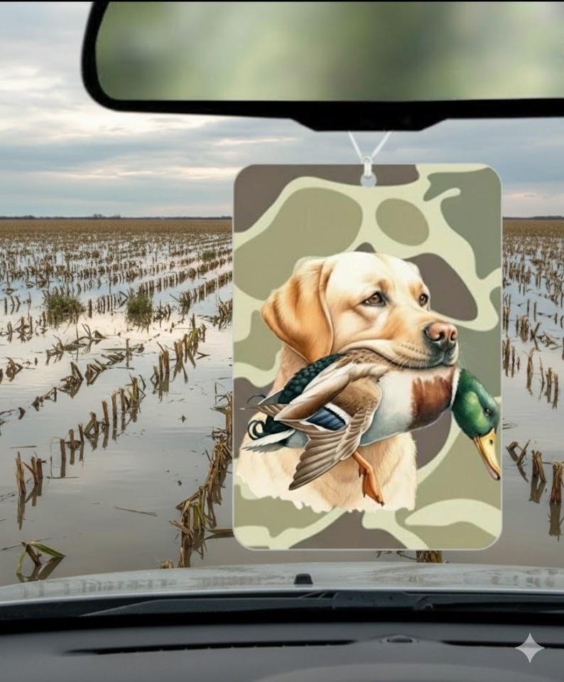 May include: Rectangular air freshener featuring a yellow Labrador Retriever holding a duck in its mouth. The background is a camouflage pattern. The air freshener hangs from a white string, with a field and sky visible in the background.