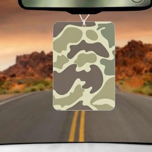 May include: Rectangular air freshener with a camouflage pattern in shades of brown, tan, and green, hanging from a car's rearview mirror. The background shows a road leading towards a sunset over a mountain range.