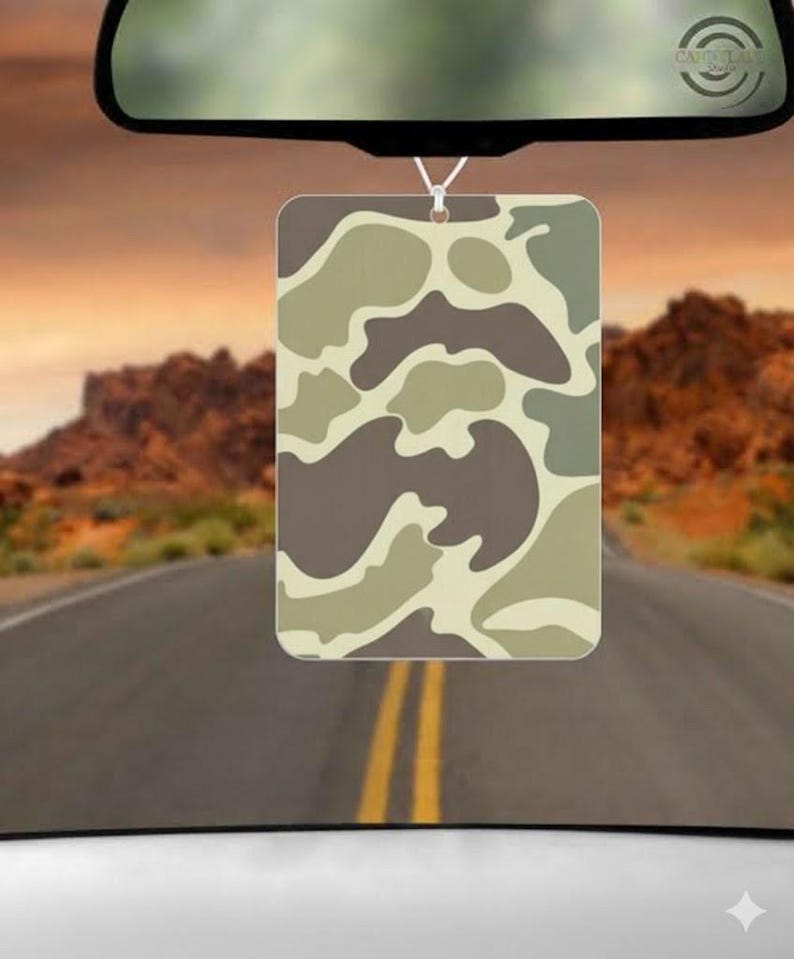 May include: Rectangular air freshener with a camouflage pattern in shades of brown, tan, and green, hanging from a car's rearview mirror. The background shows a road leading towards a sunset over a mountain range.