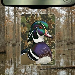 May include: A rectangular air freshener featuring a detailed illustration of a Wood Duck. The duck has iridescent green, blue, and purple feathers, a red eye, and a white and black pattern. The background is a camouflage pattern.
