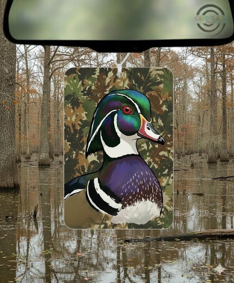 May include: A rectangular air freshener featuring a detailed illustration of a Wood Duck. The duck has iridescent green, blue, and purple feathers, a red eye, and a white and black pattern. The background is a camouflage pattern.