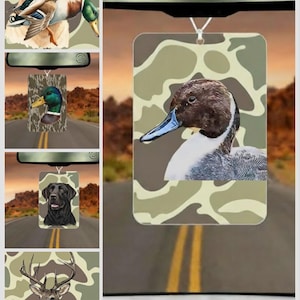 Pode incluir: Ambientador com uma ilustra&ccedil;&atilde;o de pato sobre um fundo de camuflagem. O pato tem cabe&ccedil;a castanha, bico azul e peito branco. Outros ambientadores na imagem apresentam um c&atilde;o, um veado e um pato.
