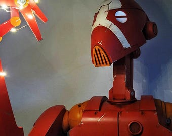 May include: A red and orange robot with a white design on its head and chest. The robot has a long neck and a segmented arm holding a string of small lights. The robot's design includes a vent-like feature on its face. The logo "Droid Forge" is visible.