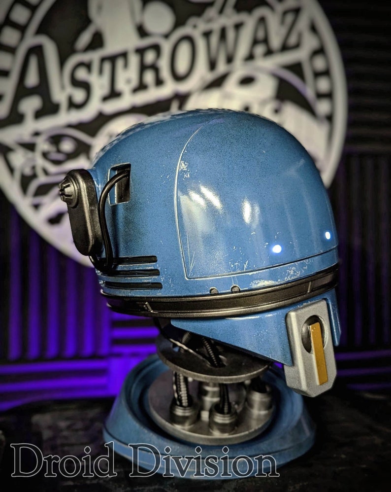 May include: A blue droid head with a metallic finish, featuring a black horizontal band, a silver and gold rectangular faceplate, and two small glowing white lights. The droid is mounted on a circular base with visible internal wiring.