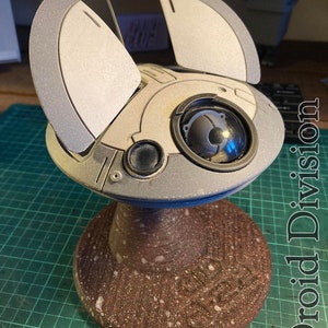 May include: A white and grey model of a futuristic drone with two large wings on the top. The drone is sitting on a brown base with white speckles. The base has the words "Droid Division" printed on the side.