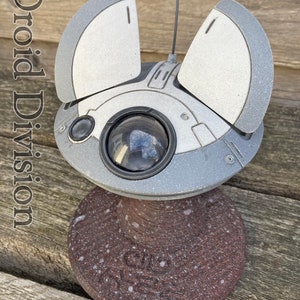 May include: A white and silver model of a Star Wars droid with a round body and two wings. The droid is sitting on a brown base with white speckles. The text "Droid Division" is visible in the image.