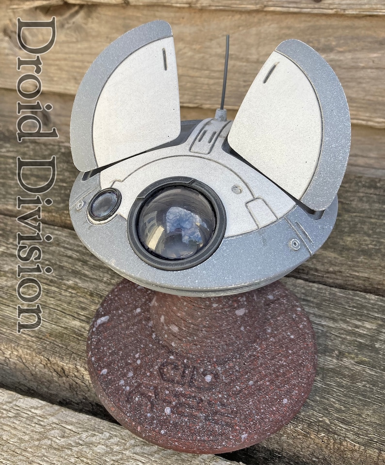 May include: A white and silver model of a Star Wars droid with a round body and two wings. The droid is sitting on a brown base with white speckles. The text "Droid Division" is visible in the image.