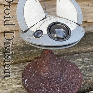 May include: A white and grey model of a futuristic spaceship with a round, black eye-like sensor. The spaceship is on a brown, speckled base. The text "Droid Division" is visible in the image.