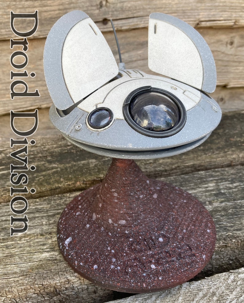 May include: A white and grey model of a futuristic spaceship with a round, black eye-like sensor. The spaceship is on a brown, speckled base. The text "Droid Division" is visible in the image.