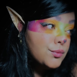 May include: A person with long black hair and pointed ears has colourful makeup on their face. The makeup includes a yellow, green, and pink design on their forehead and eye area. A small red heart is painted on their nose.