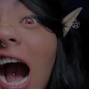 May include: A close-up of a person's face with a piercing in their nose, lip, and tongue. They are wearing a gold chain nose ring and have a red contact lens in their right eye. They have a pointed ear with a pentagram earring.