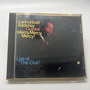 May include: A CD case featuring "The Cannonball Adderley Quintet" album, "Mercy, Mercy, Mercy!" The cover has a black background with the album title in yellow and the text "Live at The Club" in blue. A portrait of a man is visible.