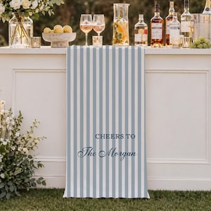 May include: A white outdoor bar with a blue and white striped front panel. The bar is decorated with floral arrangements, glassware, and bottles of spirits. The panel reads "Cheers to The Morgan" in navy blue script.