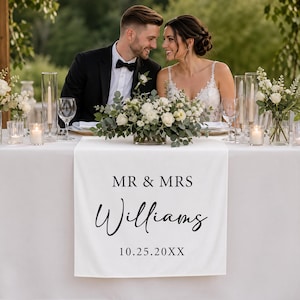 Puede incluir: Un camino de mesa blanco con el texto "MR & MRS Williams" y la fecha "10.25.20XX" en escritura negra. La mesa está puesta para una boda, con arreglos florales, velas y cristalería. Una pareja está sentada a la mesa.