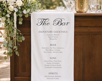 May include: A wooden bar with a white cloth displaying "The Bar" in elegant script. The bar offers signature cocktails, beer, wine, and spirits. A floral arrangement and glassware are visible.