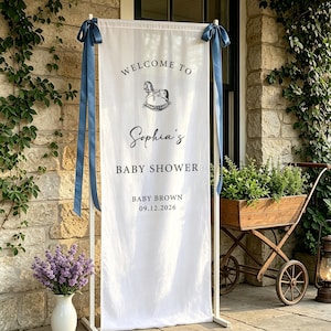 Puede incluir: Una pancarta blanca con lazos azules, que muestra "WELCOME TO Sophia's BABY SHOWER" y un gr&aacute;fico de caballito balanc&iacute;n. La pancarta est&aacute; sostenida por un marco de metal blanco. La fecha "09.12.2026" tambi&eacute;n est&aacute; impresa.