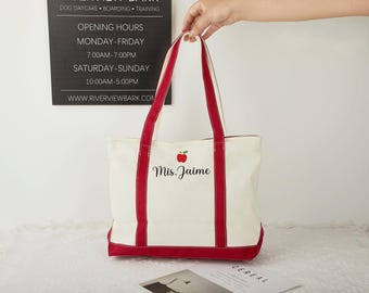 May include: A white canvas tote bag with red trim and a red apple graphic. The bag has the text "Mis. Jaime" embroidered on the front.