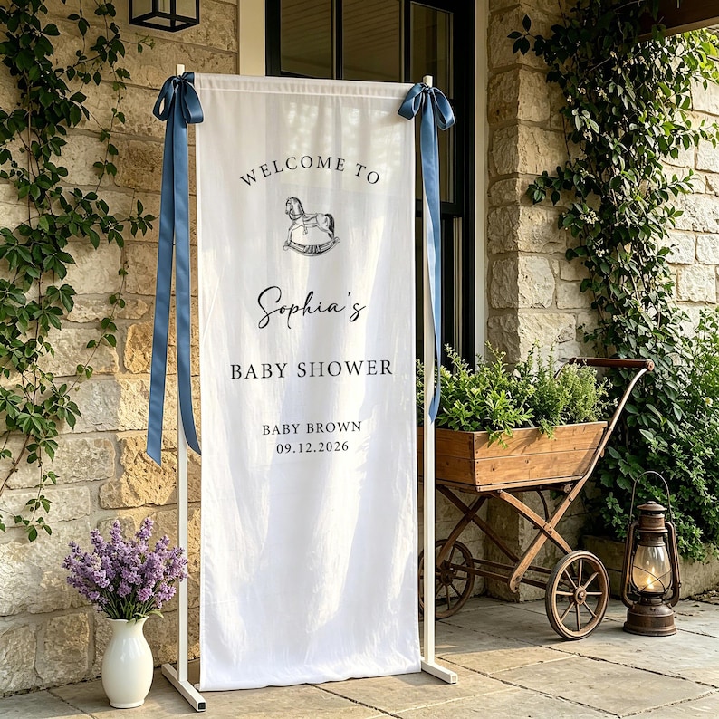 Puede incluir: Una pancarta blanca con lazos azules, que muestra "WELCOME TO Sophia's BABY SHOWER" y un gr&aacute;fico de caballito balanc&iacute;n. La pancarta est&aacute; sostenida por un marco de metal blanco. La fecha "09.12.2026" tambi&eacute;n est&aacute; impresa.
