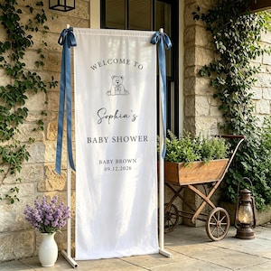 Puede incluir: Una pancarta blanca de baby shower con el texto "Welcome to Sophia's Baby Shower" y la fecha 09.12.2026. La pancarta est&aacute; sujeta por un marco de metal blanco y adornada con cintas azules. Un dibujo de un oso de peluche est&aacute; en la parte superior.