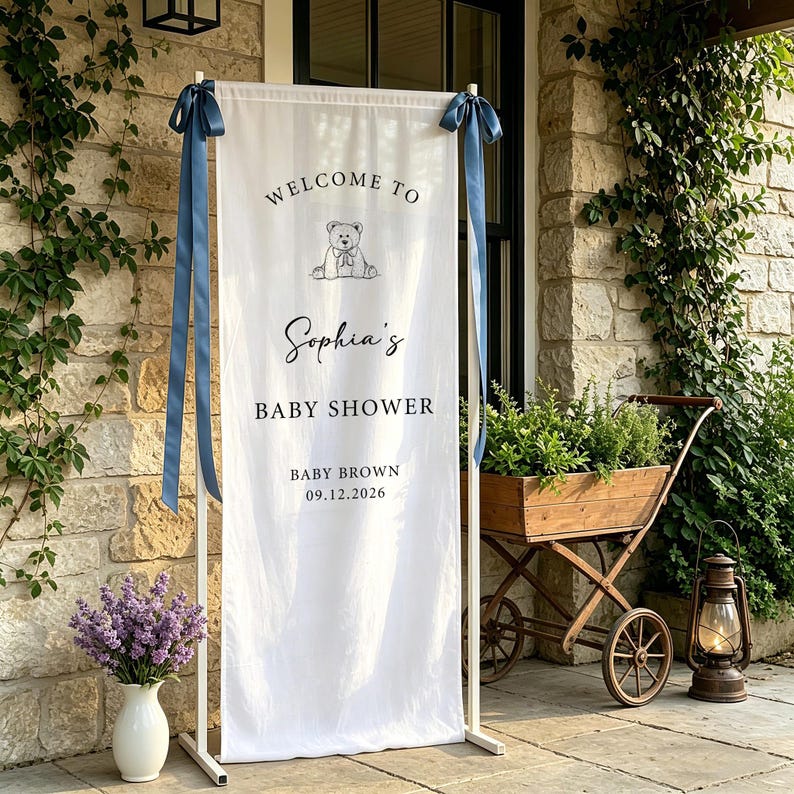 Puede incluir: Una pancarta blanca de baby shower con el texto "Welcome to Sophia's Baby Shower" y la fecha 09.12.2026. La pancarta est&aacute; sujeta por un marco de metal blanco y adornada con cintas azules. Un dibujo de un oso de peluche est&aacute; en la parte superior.