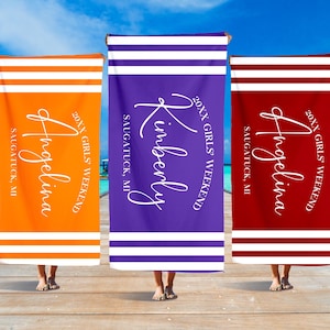 Op de afbeelding: Drie strandhanddoeken met witte strepen en de tekst "20XX Girls' Weekend" en een naam op elke handdoek. De handdoeken zijn oranje, paars en rood.