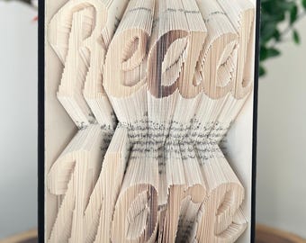 May include: A book art sculpture with the words "Read More" folded into the pages. The book is bound in black with gold accents and is sitting on a wooden surface.