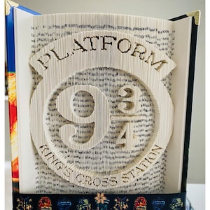 Peut inclure: Sculpture de livre avec les mots "PLATFORM 9 3/4" et "KINGS CROSS STATION" découpés dans les pages. Le livre ouvert révèle le design complexe. Il a un dos bleu et des coins dorés. Un ruban décoratif est en bas.