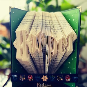 Könnte beinhalten: Ein Buch, das in das Wort "Harry" geformt ist, mit einem grünen Einband mit weißen Sternen und einem schwarzen Band mit bunten Symbolen. Das Band trägt die Aufschrift "Bookgami by Toppy".
