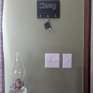 May include: A black rectangular wooden wall-mounted key holder featuring the word Chevy in white lettering. The holder has three silver metal hooks, with a set of car keys hanging from the center hook. The item is mounted on a sage green wall.