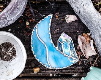 May include: A blue and iridescent glass suncatcher shaped like a crescent moon with a crystal-like design. The suncatcher is hanging from a black ribbon and is resting on a wooden surface.