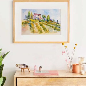 Op de afbeelding: Aquarel schilderij van een Toscaans landschap met een wijngaard, bomen en gebouwen, omlijst in licht hout. Het kunstwerk hangt boven een houten dressoir met decoratieve items.