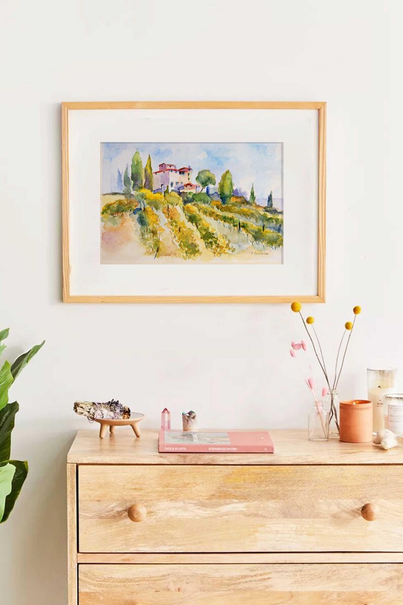 Op de afbeelding: Aquarel schilderij van een Toscaans landschap met een wijngaard, bomen en gebouwen, omlijst in licht hout. Het kunstwerk hangt boven een houten dressoir met decoratieve items.
