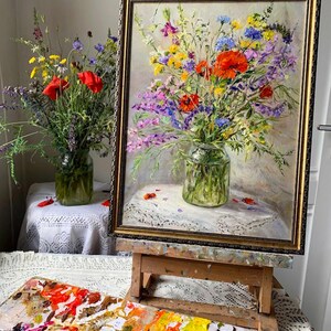 May include: An oil painting of a vibrant floral arrangement in a glass vase, framed in gold. The bouquet features red poppies, purple, yellow, and blue wildflowers. A wooden easel supports the artwork, with a palette of paint colors in the foreground.