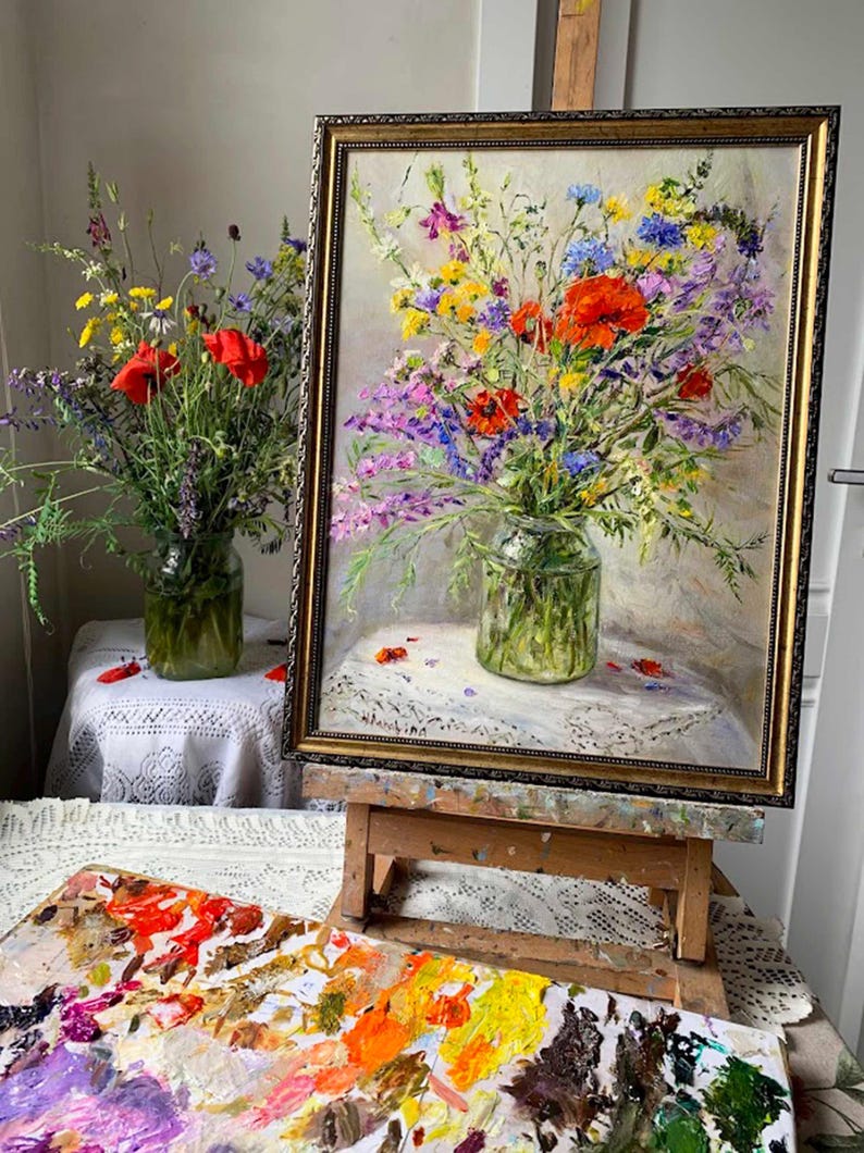 May include: An oil painting of a vibrant floral arrangement in a glass vase, framed in gold. The bouquet features red poppies, purple, yellow, and blue wildflowers. A wooden easel supports the artwork, with a palette of paint colors in the foreground.