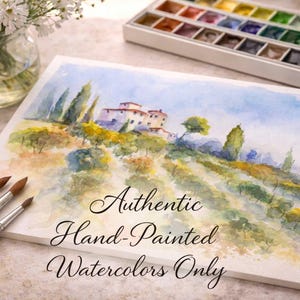 Op de afbeelding: Een aquarel van een Toscaans landschap met heuvels, bomen en een gebouw. De schildering staat op een wit canvas met de tekst "Authentic Hand-Painted Watercolors Only." Penselen en een aquarelpalet zijn ook zichtbaar.