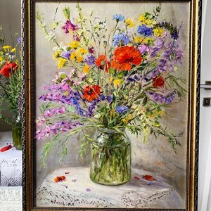 May include: An oil painting of a vibrant bouquet of wildflowers in a clear glass vase. The flowers include red poppies, purple, yellow, and blue blooms. The painting is in a gold ornate frame and rests on a white lace tablecloth.