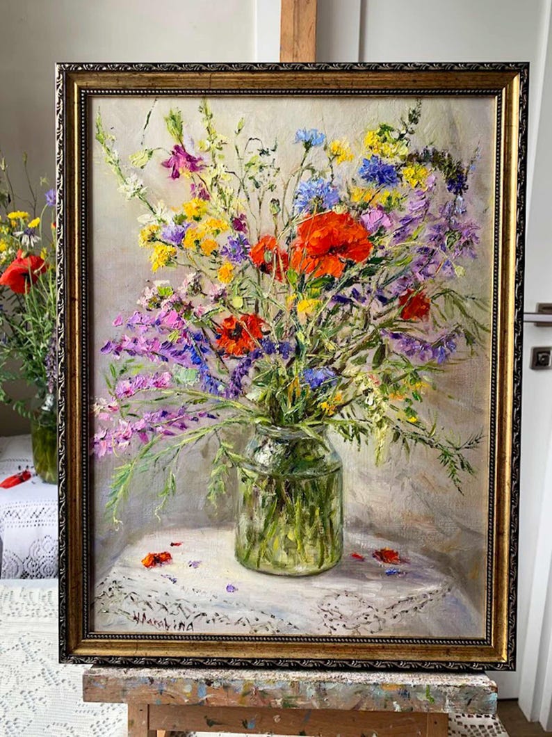 May include: An oil painting of a vibrant bouquet of wildflowers in a clear glass vase. The flowers include red poppies, purple, yellow, and blue blooms. The painting is in a gold ornate frame and rests on a white lace tablecloth.