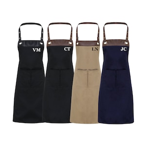 Puede incluir: Cuatro delantales negros, marrones, beige y azul marino con correas de cuero marrón y ojales metálicos. Cada delantal tiene un bolsillo y un monograma bordado en blanco o negro: VM, CT, LN y JC.