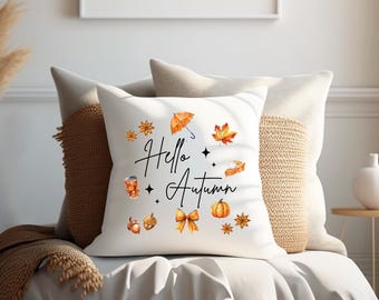 Puede incluir: Almohada blanca con el texto "Hello Autumn" en letras negras. La almohada está decorada con ilustraciones de temática otoñal, que incluyen una calabaza, hojas, un paraguas y una taza de café.