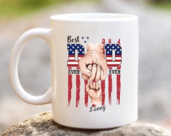 May include: White ceramic coffee mug with a design of two fists bumping, one larger and one smaller, with the word "DAD" in large letters and the words "Best Ever" on either side. The design is set against a red, white, and blue American flag background. The name "Lenny" is printed at the bottom of the mug.