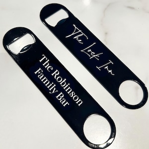 Op de afbeelding: Twee zwarte metalen flessenopeners met witte tekst. De bovenste opener zegt "The Lock Inn" en de onderste opener zegt "The Robinson Family Bar".