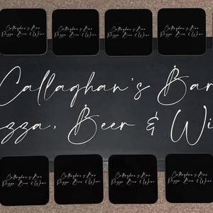 May include: Black coasters with white text that reads "Callaghan's Bar Pizza, Beer & Wine". The coasters are arranged in two rows on a black surface.