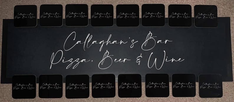 May include: Black coasters with white text that reads "Callaghan's Bar Pizza, Beer & Wine". The coasters are arranged in two rows on a black surface.