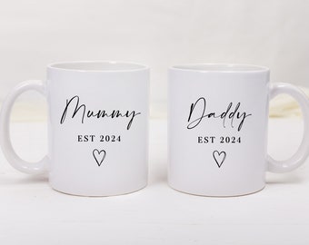 May include: Two white ceramic mugs with black lettering. One mug says "Mummy EST 2024" with a small heart. The other mug says "Daddy EST 2024" with a small heart.
