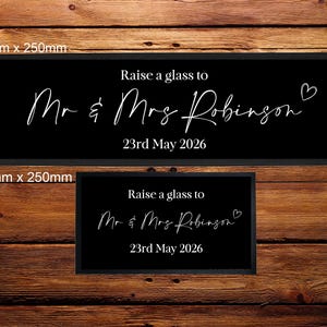 May include: Two black rectangular signs with white text on a wooden background. The larger sign reads "Raise a glass to Mr & Mrs Robinson 23rd May 2026". The smaller sign has the same text. The signs are 880mm x 250mm and 440mm x 250mm.