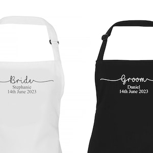 以下が含まれることがあります： 「Bride」と「Groom」の文字が印刷された黒と白の2つのエプロン。花嫁のエプロンは黒文字で白、新郎のエプロンは白文字で黒。両方のエプロンには「2023年6月14日」の文字が印刷されています。