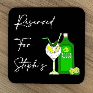 Op de afbeelding: Zwarte onderzetter met witte tekst die "Gereserveerd voor Steph's" zegt. Een groene gin-fles met een kroon en het woord "GIN" staat naast een glas gin met een limoenpartje en een rietje.