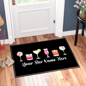 May include: Black welcome mat with a white border and the text "Your Bar Name Here". The mat features a row of six colourful cocktail glasses with various garnishes.