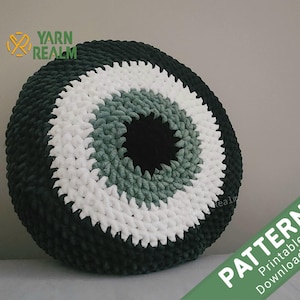 Könnte beinhalten: Ein rundes gehäkeltes Kissen mit einem großen Augenmotiv in Grün-, Weiß- und Schwarztönen. Das Kissen ist aus dickem Garn gefertigt und hat eine strukturierte Oberfläche. Der Text "PATTERN Printable Download File" ist in der unteren rechten Ecke des Bildes sichtbar.